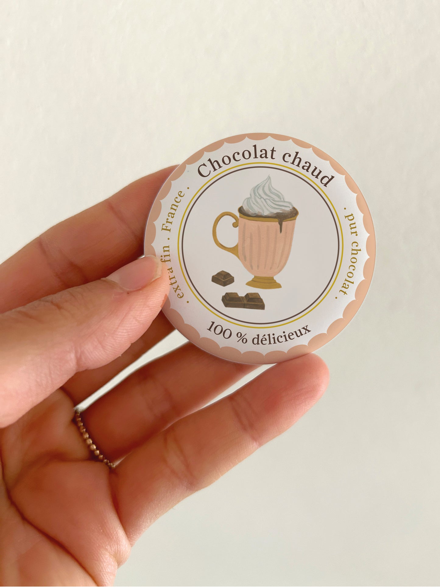 Ein runder 55mm Kühlschrankmagnet mit mattem, reflexionsfreiem Finish, gehalten in einer Hand. Das handgemalte Aquarell-Design zeigt eine edle Schokoladenpraline, umgeben von einem braunen Wellenrand