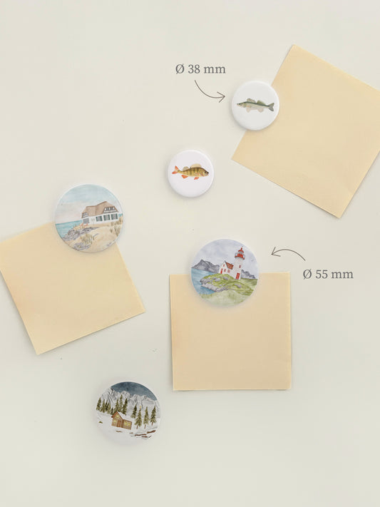 Magnetwand mit verschiedenen, runden Magneten, die handillustrierter Häuser in einer schönen Landschaft zeigen, zum Beispiel ein Strandhaus, einen Leuchtturm oder einen Holzhütte im Schnee.