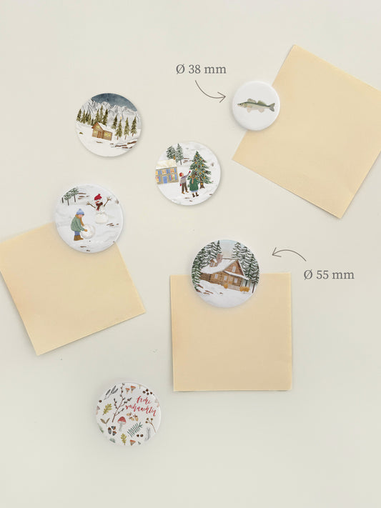 Magnetwand mit verschiedenen, runden Magnetbuttons mit weihnachtlichen Motiven, wie zum Beispiel Holzhütten im Winterwald, Kinder beim Baumschmücken oder Schneemann bauen oder Naturcollagen mit "Fröhliche Weihnachten"-Aufschrift.