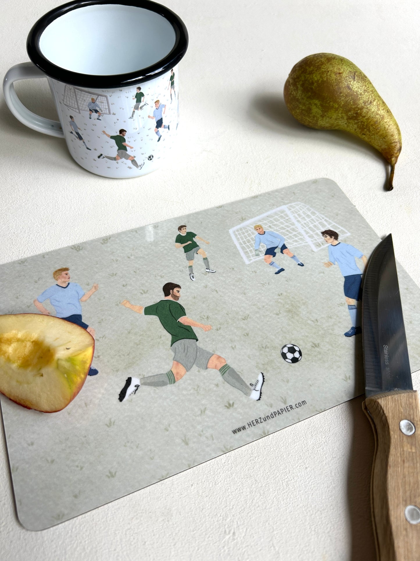 Ein graugrünes Frühstücksbrettchen mit handgemalten Illustrationen spielender Fußballer liegt auf einer hellen Tischplatte. Im Vordergrund schneidet ein Holzmesser ein Stück Apfel direkt auf dem Brettchen an. Links oben steht eine weiße Emaille-Tasse mit schwarzem Rand und passendem Fußball-Motiv, rechts daneben liegt eine frische Birne. Am unteren Rand des Brettchens ist die dezente Webadresse „www.HERZundPAPIER.com“ zu sehen.