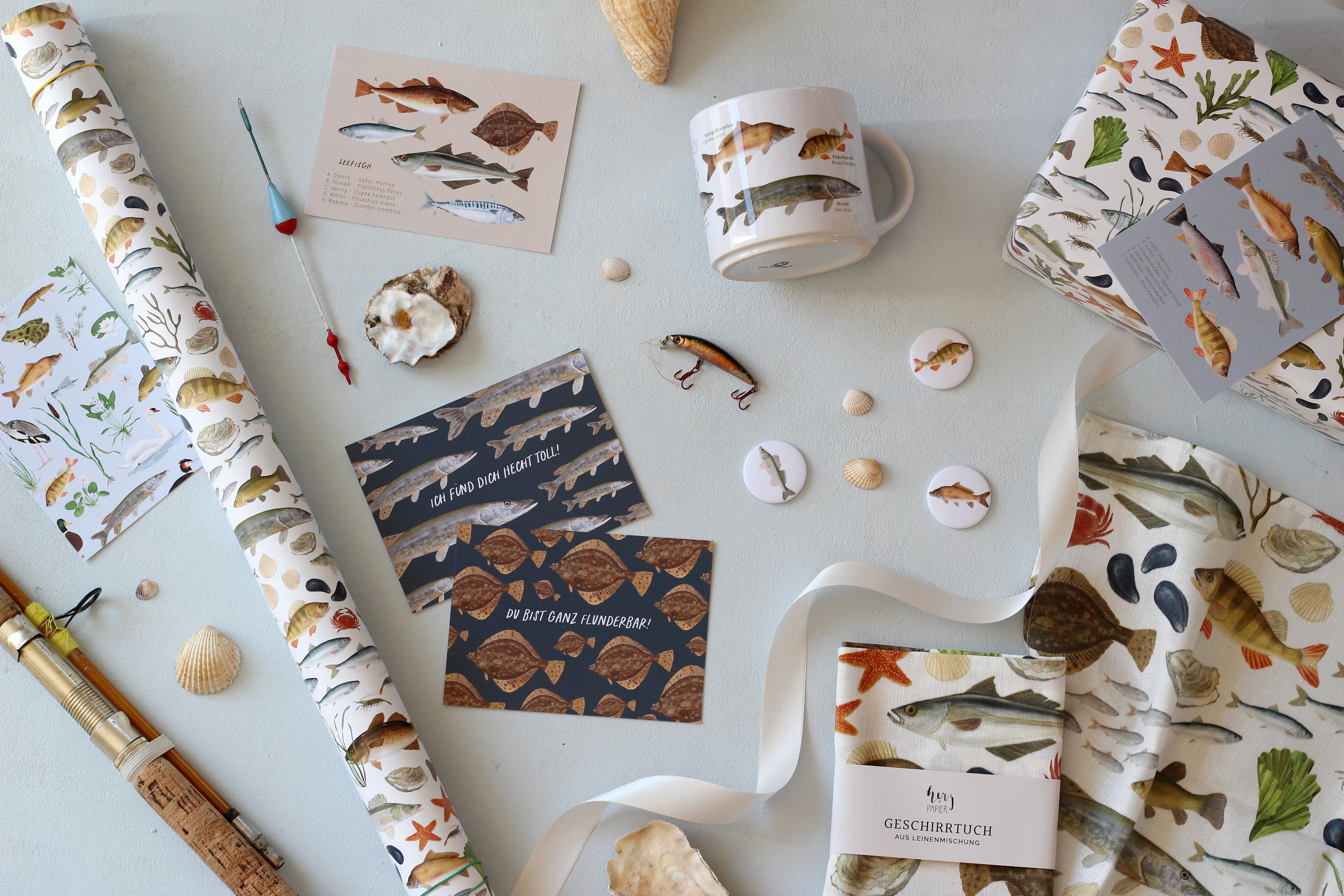 Flatlay mit verschiedenen Produkte mit Fisch-Illustrationen. Zum Beispiel Geschenkpapier, Postkarten, Magnete, Tassen, Geschirrtuch. Alle Produkte zieren handgemalte, realistische Illustrationen von heimischen Fischen wie Hecht, Forelle, Zander, Karpfen und Seefisch. 