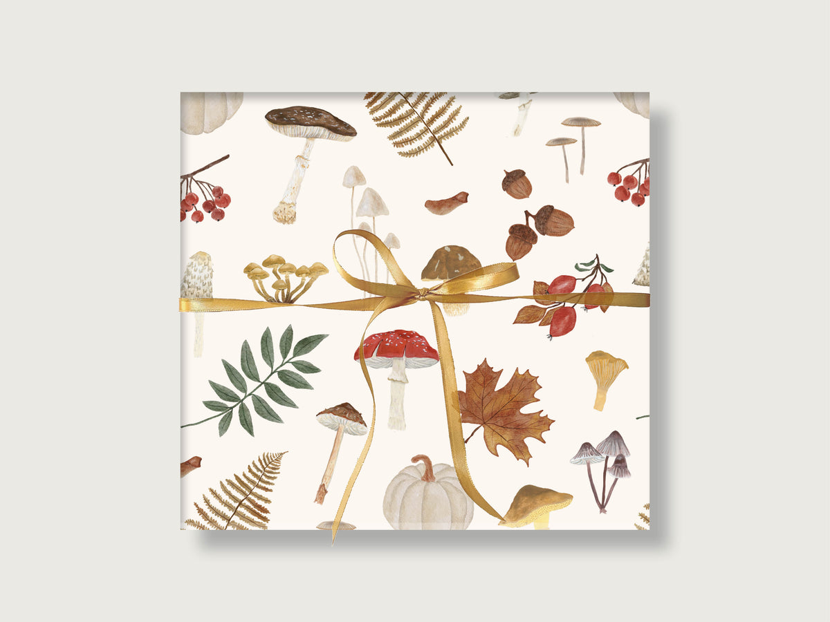 Eingepacktes Geschenk mit mattem Geschenkpapier im herbstlichen Waldmotiv aus Pilzen, Eicheln, Farnen und hellem Kürbis, gebunden mit einer goldenen Schleife.