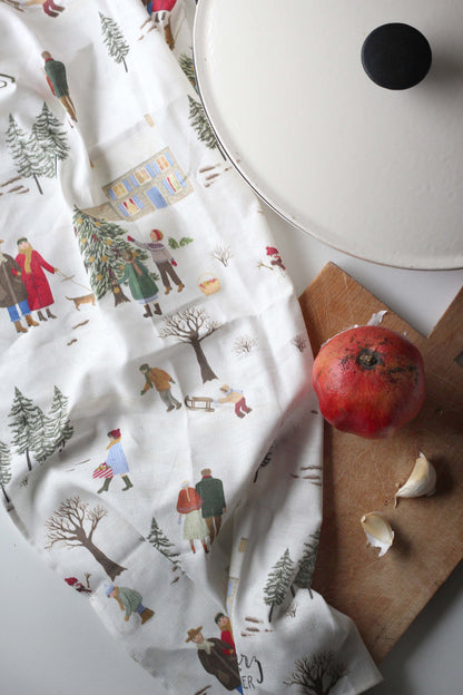 Geschirrtuch aus Halbleinen mit handillustriertem Winterdorf, Tannenbaum und Schlittenfahrern neben einer roten Gusseisenpfanne auf der Küchenzeile.   