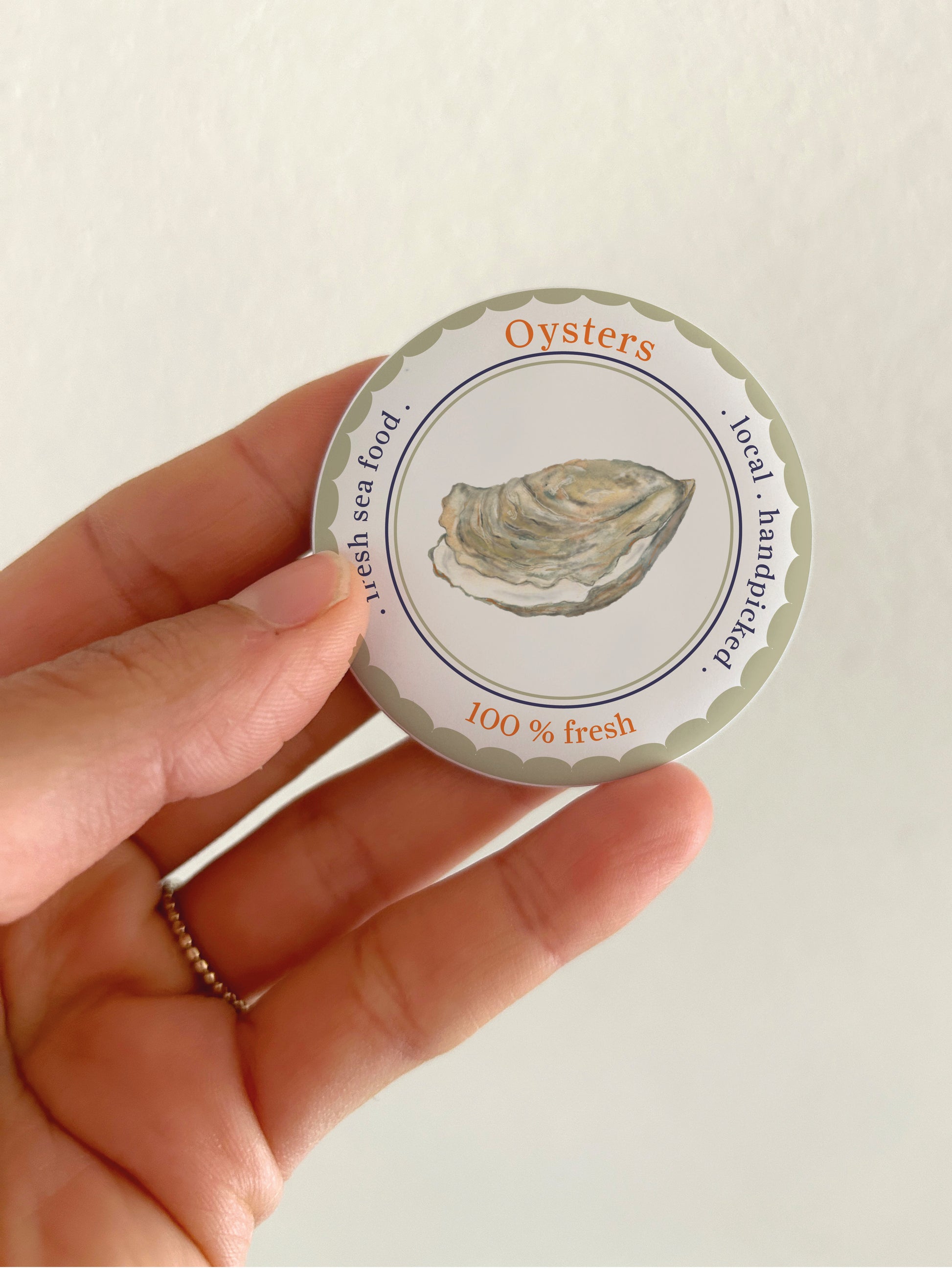 Ein runder 55mm Kühlschrankmagnet mit matter Oberfläche, der in einer Hand gehalten wird. Das handgemalte Aquarell-Motiv zeigt eine geöffnete Auster mit feinen Details in Blau- und Grautönen. Ein dunkelblauer Wellenrand und ein Schriftzug runden das maritime Design ab.