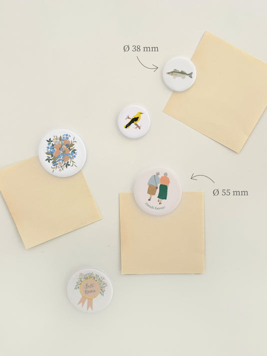 Magnetwand mit verschiedenen runden Magnet-Buttons mit handillustrierten Motiven. Zu sehen sind Motive mit Blumen, besten Freunden und für die beste Mama. Die Magnete sind weiß und rund mit Motiv.