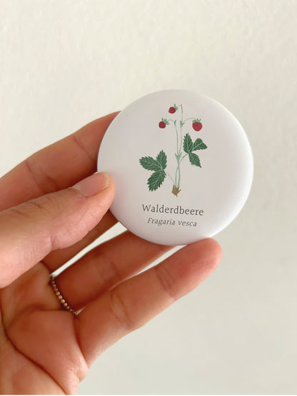 Ein runder 55mm Kühlschrankmagnet mit mattem Finish, der in einer Hand gehalten wird. Das handgemalte Aquarell-Motiv zeigt eine filigrane Walderdbeere mit grünen Blättern und drei kleinen, roten Beeren samt der Aufschrift „Walderdbeere – Fragaria vesca“.
