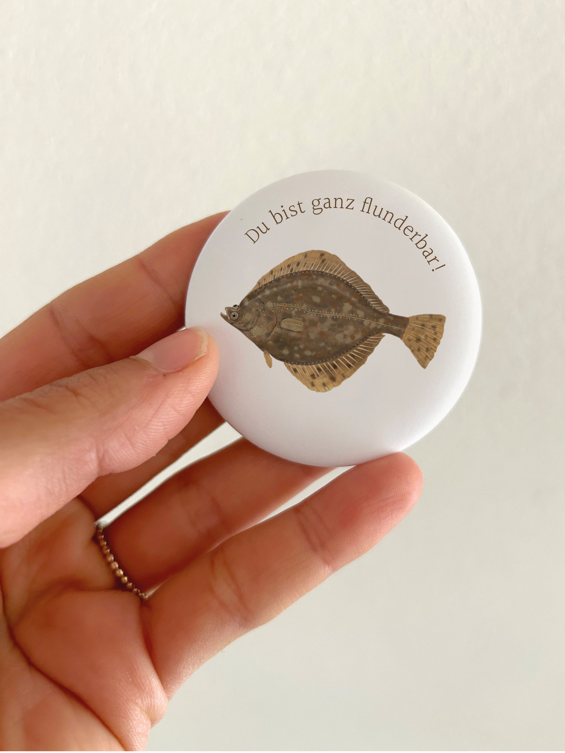 Ein runder, matter Kühlschrankmagnet (55mm Durchmesser), der in einer Hand gehalten wird. Das Motiv zeigt den hochwertigen Druck einer detailreichen, handgemalten Flunder auf weißem Grund, abgerundet durch den geschwungenen, braunen Schriftzug „Du bist ganz flunderbar!“.