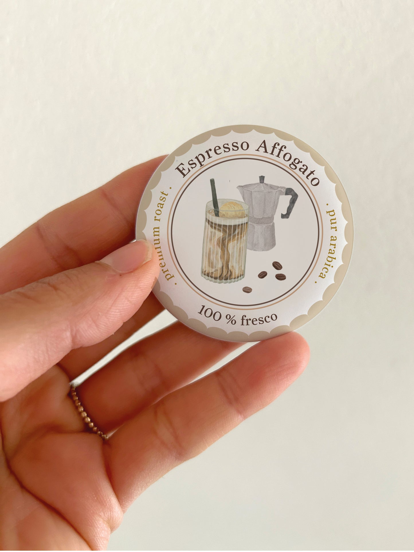 Ein runder, matter 55mm Kühlschrankmagnet in einer Hand. Das handgemalte Aquarell-Motiv zeigt eine klassische Espressokanne (Mokkakanne) in Grau- und Blautönen, umrahmt von einem braunen Wellenrand