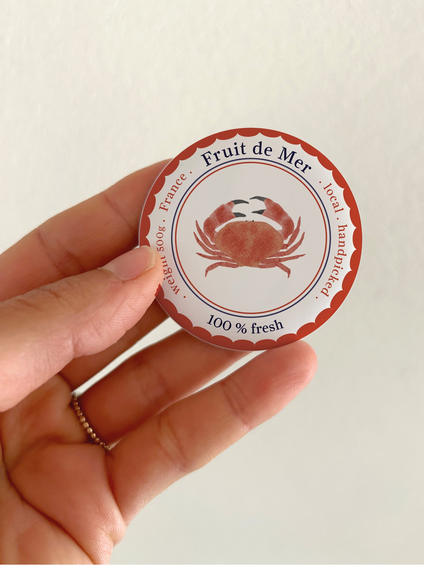 Ein runder 55mm Kühlschrankmagnet mit mattem Finish, präsentiert in einer Hand. Das handgemalte Aquarell-Motiv zeigt eine rötlich-braune Krabbe auf weißem Grund, verziert mit einem blauen Wellenrand