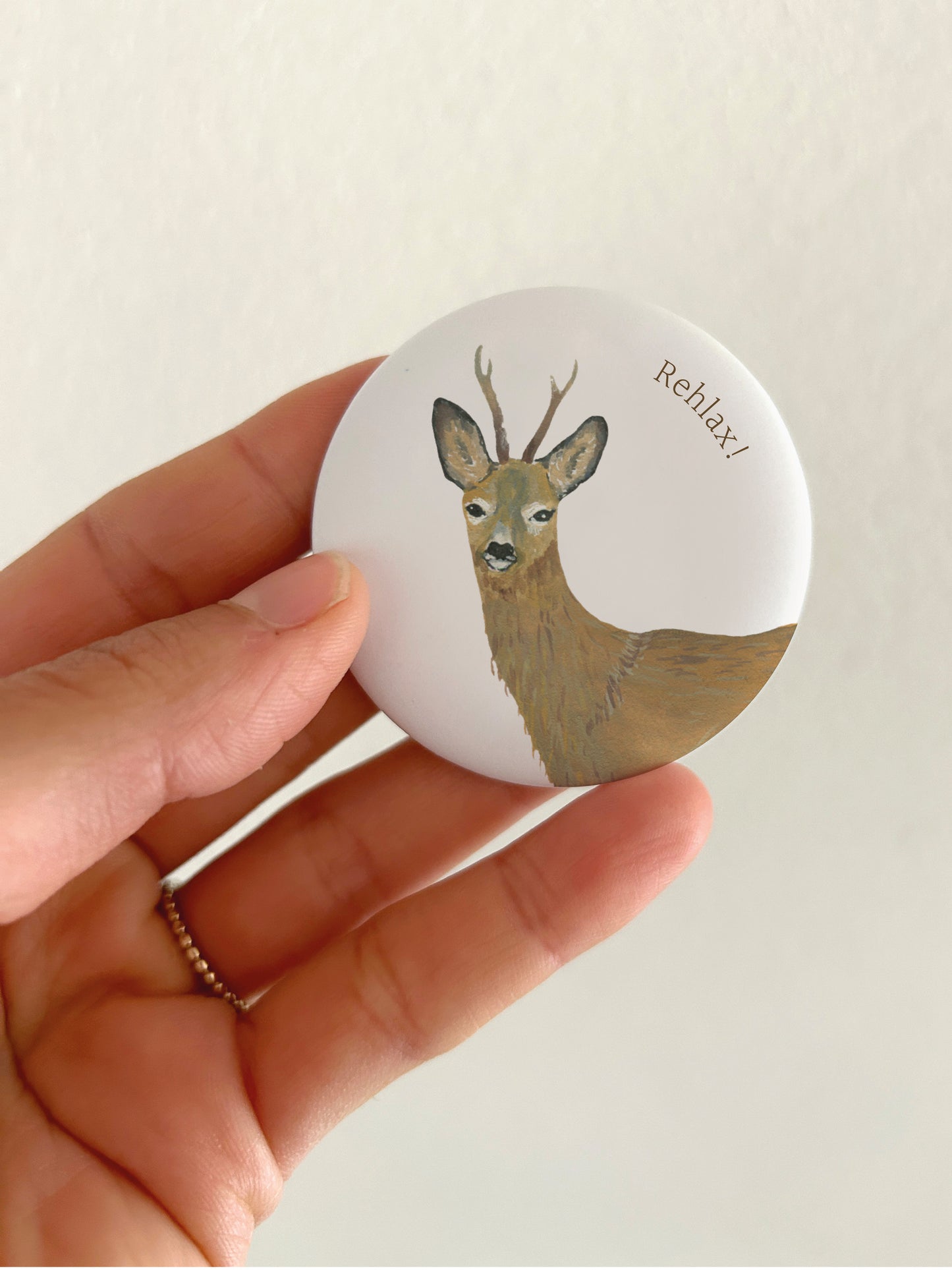 Ein runder, matter 55mm Kühlschrankmagnet, der in einer Hand gehalten wird. Zu sehen ist der hochwertige Druck einer detailreichen, handgemalten Aquarell-Illustration eines Rehbocks mit Geweih auf weißem Grund. Oben rechts steht in feiner, brauner Schrift das Wortspiel „Rehlax!“.