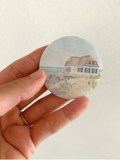 Großer maritimer Strandhaus-Kühlschrankmagnet mit 55mm Durchmesser als Zubehör und Deko für das Wohnmobil oder Vanlife. Eine Hand präsentiert den runden, matten Magneten vor einer hellen Wand, um das reflexionsfreie Oberflächenfinish, das Reetdach-Motiv und die reale Produktgröße zu zeigen