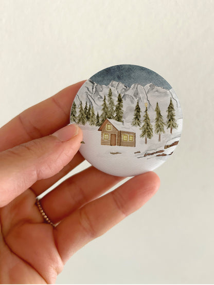 Großer Weihnachts-Magnet mit verschneiter Berghütte und 55mm Durchmesser als winterliche Dekoration für die Kühlschranktür. Ein runder, matter Magnet mit handgemalter Natur-Illustration wird als Größenvergleich vor einem hellen Hintergrund in einer Hand gehalten, um die matte, kratzfeste und spiegelungsfreie Oberfläche zu präsentieren.