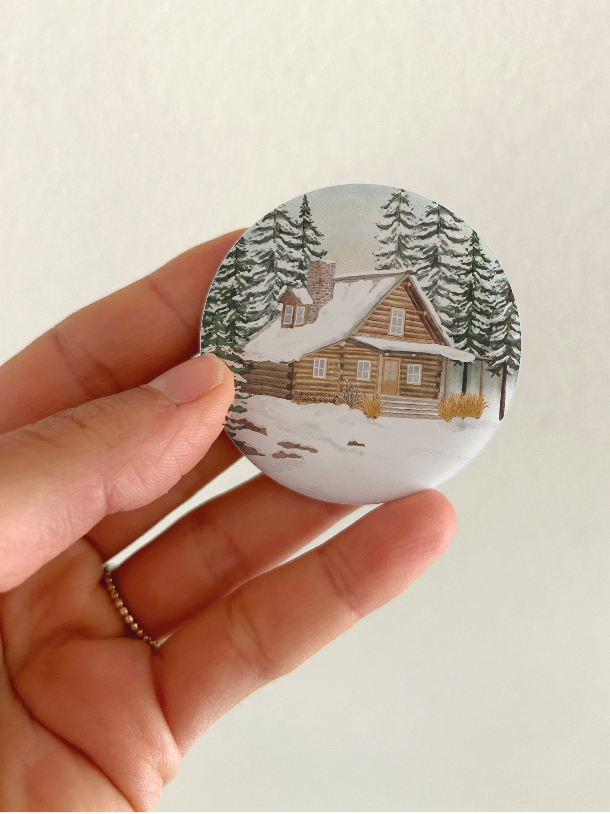 Großer Weihnachts-Magnet mit verschneiter Blockhütte und 55mm Durchmesser als winterliche Dekoration für die Kühlschranktür. Ein runder, matter Magnet mit handgemalter Natur-Illustration aus Große-Magnete-Weihnachten2.jpg wird als Größenvergleich vor einem hellen Hintergrund in einer Hand gehalten, um die matte, kratzfeste und spiegelungsfreie Oberfläche zu präsentieren.
