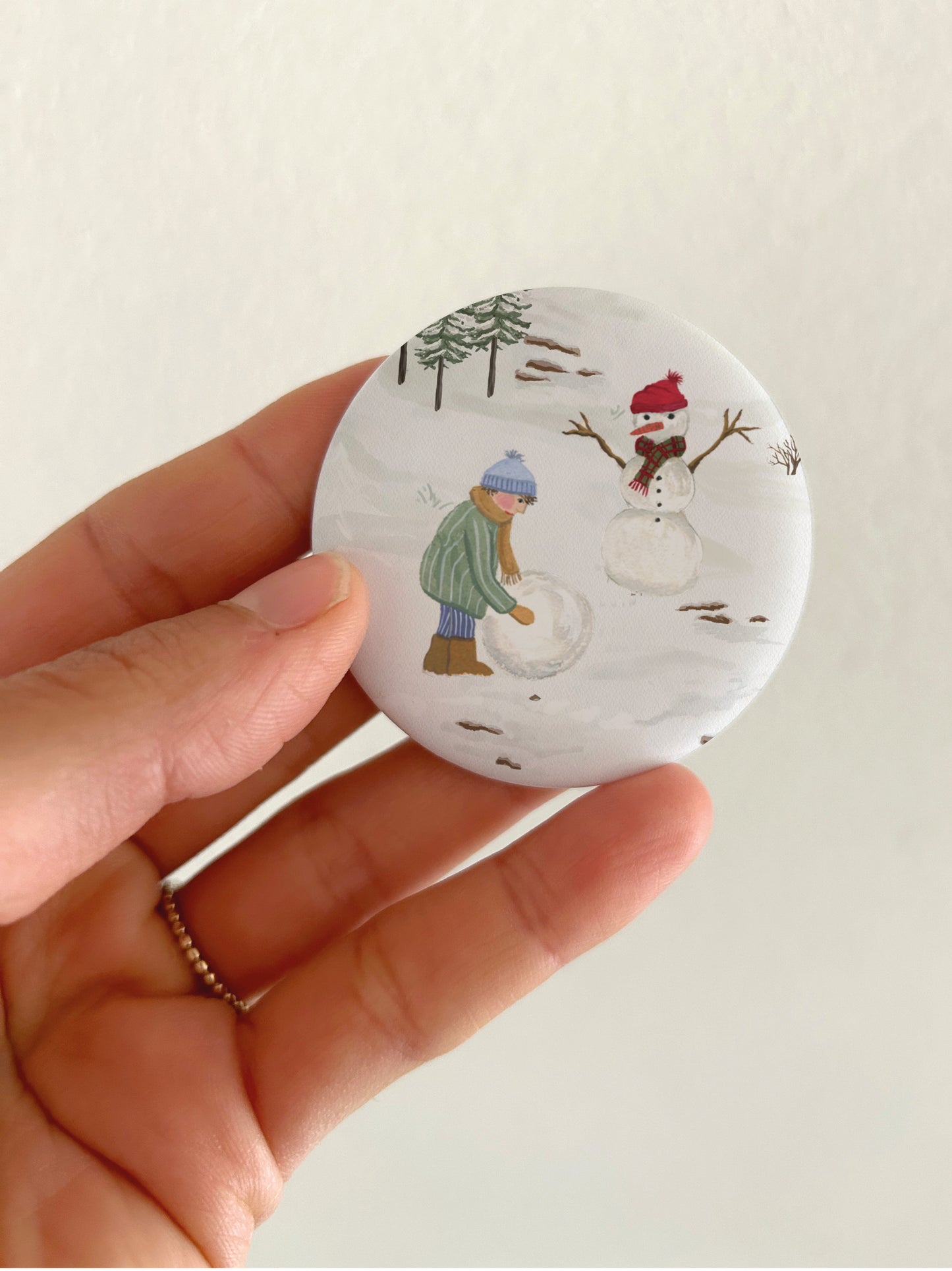 Großer Weihnachts-Magnet mit winterlicher Schneemann-Szene und 55mm Durchmesser als Dekoration für die Kühlschranktür. Ein runder, matter Magnet mit handgemalter Natur-Illustration wird als Größenvergleich vor einem hellen Hintergrund in einer Hand gehalten, um die matte, kratzfeste und spiegelungsfreie Oberfläche zu präsentieren.