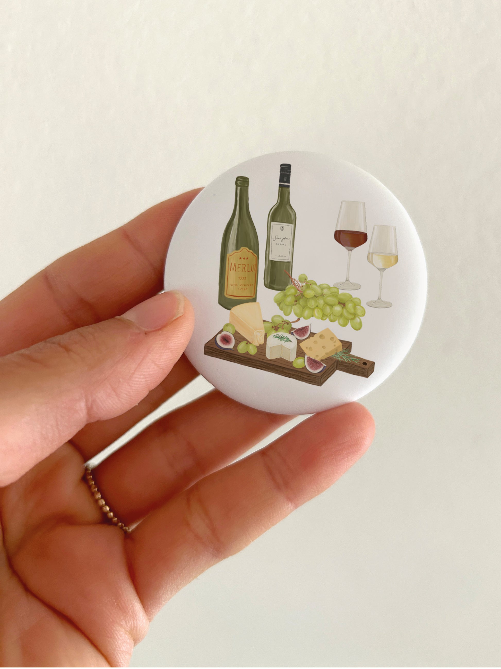 Eine Hand hält einen runden, weißen Kühlschrankmagnet, auf dem ein handgemaltes Wein und Käse-Motiv zu sehen ist. Neben einer Flasche Rotwein und Weißwein sowie einem Glas Rotwein und Weißwein ist im Vordergrund eine Käseplatte und Trauben zu sehen. 