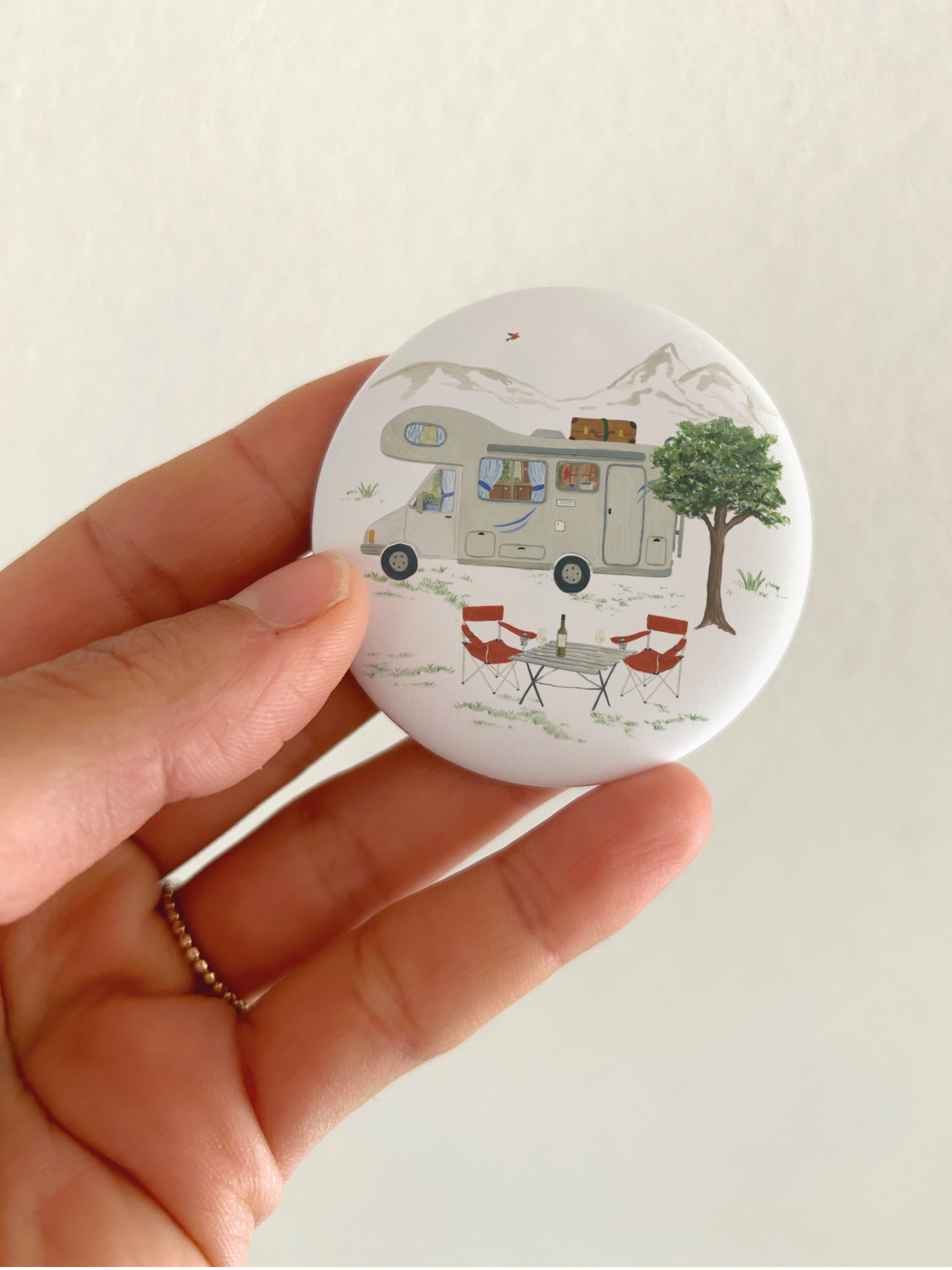 Kühlschrankmagnet mit Camping-Motiv, das in der HAnd gehalten wird. Neben dem Wohnmobil sieht man auch Campingausrüstung und Landschaft.