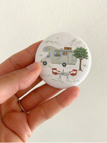 Kühlschrankmagnet mit Camping-Motiv, das in der HAnd gehalten wird. Neben dem Wohnmobil sieht man auch Campingausrüstung und Landschaft.