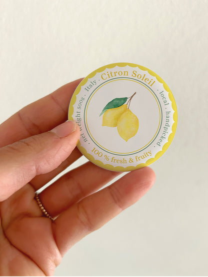 Ein runder Kühlschrankmagnet (55mm) mit mattem Finish, gehalten in einer Hand. Das handgemalte Aquarell-Motiv im mediterranen Stil zeigt zwei gelbe Zitronen mit grünen Blättern. Umrahmt ist das Design von einem gelben Wellenrand und der eleganten Aufschrift „Citron Soleil - Italy - 100% fresh & fruity“.