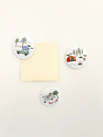 3 runde Magnete mit Campingmotiven an einer Magnetwand. Die großen Magnete Buttons zeigen Wohnmobil, Zelt und Jeep mit Dachzelt 