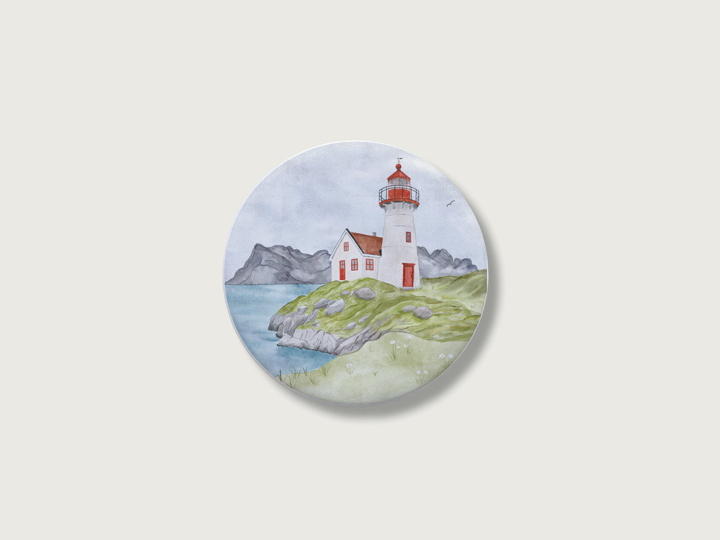 Maritimes Geschenk für Küstenkinder und Camper: Runder Kühlschrankmagnet mit einer malerischen, detailreichen Handillustration eines klassischen rot-weißen Leuchtturms und Wärterhäuschens auf einer grünen, felsigen Klippe direkt am Meer vor einer weiten Bergkulisse im Hintergrund.