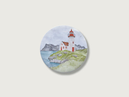 Maritimes Geschenk für Küstenkinder und Camper: Runder Kühlschrankmagnet mit einer malerischen, detailreichen Handillustration eines klassischen rot-weißen Leuchtturms und Wärterhäuschens auf einer grünen, felsigen Klippe direkt am Meer vor einer weiten Bergkulisse im Hintergrund.