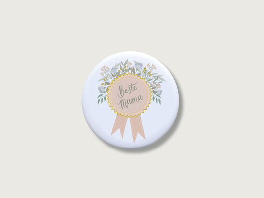 Liebevolles Geschenk zum Muttertag oder Geburtstag: Runder, weißer Kühlschrankmagnet in Form einer Ehrenmedaille mit Schleife, einem feinen Blütenkranz in sanften Naturtönen und dem handillustrierten Schriftzug Beste Mama. Das dekorative Motiv für die Magnetwand ist mittig auf einem schlichten, neutralen, hellen Hintergrund platziert.