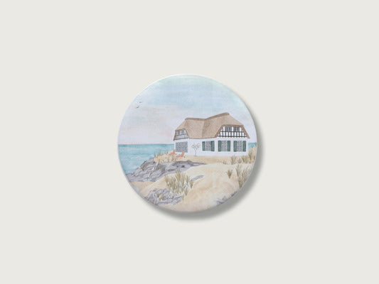Maritimes Geschenk für Küstenkinder und Camper: Runder Kühlschrankmagnet mit einer malerischen, detailreichen Handillustration eines klassischen Reetdachhauses mit grünen Fensterläden in einer Dünenlandschaft mit Strandhafer direkt am weiten, hellblauen Meer.