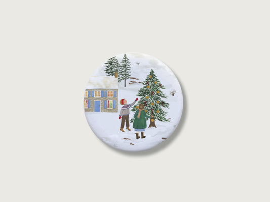 Nostalgisches Geschenk zur Adventszeit und für Familien: Runder, weißer Kühlschrankmagnet mit einer detailreichen, handillustrierten Winterszene, bestehend aus zwei Kindern im Schnee, die eine Tanne mit Kugeln und Schleifen vor einem zweistöckigen Steinhaus mit blauen Fensterläden schmücken. Das weihnachtliche Motiv befindet sich zentriert auf einem schlichten, neutralen, hellen Hintergrund.