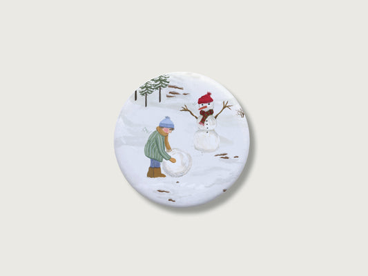 Nostalgisches Geschenk zur Adventszeit und für Familien: Runder, weißer Kühlschrankmagnet mit einer detailreichen, handillustrierten Winterszene, bestehend aus einem Kind in Winterkleidung, das eine Schneekugel rollt, und einem fertigen Schneemann mit roter Mütze und kariertem Schal. Das weihnachtliche Motiv von Großer-Magnet-Weihnachten-Schneemann.jpg befindet sich zentriert auf einem schlichten, neutralen, hellen Hintergrund.