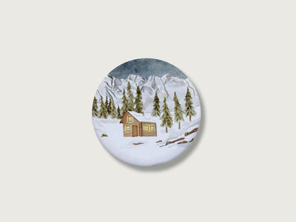 Gemütliches Geschenk zur Adventszeit und für Bergfans: Runder, weißer Kühlschrankmagnet mit einer detailreichen, handillustrierten Winterlandschaft, bestehend aus einer verschneiten Holzhütte mit leuchtenden Fenstern, tiefgrünen Tannen und einer majestätischen, schneebedeckten Bergkulisse. Das weihnachtliche Motiv von Großer-Magnet-Weihnachten1.jpg befindet sich mittig auf einem schlichten, neutralen, hellen Hintergrund.