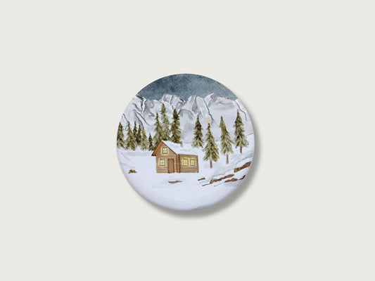 Gemütliches Geschenk zur Adventszeit und für Bergfans: Runder, weißer Kühlschrankmagnet mit einer detailreichen, handillustrierten Winterlandschaft, bestehend aus einer verschneiten Holzhütte mit leuchtenden Fenstern, tiefgrünen Tannen und einer majestätischen, schneebedeckten Bergkulisse. Das weihnachtliche Motiv von Großer-Magnet-Weihnachten1.jpg befindet sich mittig auf einem schlichten, neutralen, hellen Hintergrund.