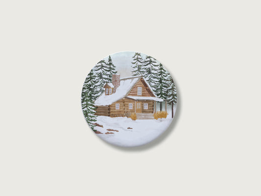 Gemütliches Geschenk zur Adventszeit und für Naturfans: Runder, weißer Kühlschrankmagnet mit einer detailreichen, handillustrierten Winterlandschaft, bestehend aus einer schneebedeckten Holzblockhütte mit gemauertem Schornstein inmitten von hohen, tief verschneiten Tannenbäumen. Das weihnachtliche Motiv befindet sich zentriert auf einem schlichten, neutralen, hellen Hintergrund.