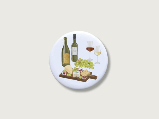 Magnet Button für Weinliebhaber mit illustriertem Wein-Motiv neben Käseplatte und Trauben. Die handgemalte Illustration assoziiert eine Weinprobe mit Weißwein und Rotwein sowie verscheidenem Käse und Trauben und Feigen. 