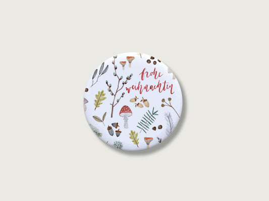 Botanische Weihnachtsdeko für die Küche: Runder, weißer Kühlschrankmagnet frohe weihnachten mit einer detailreichen, feinen Handillustration von verschiedenen Naturelementen aus dem Winterwald wie einem roten Fliegenpilz, grünen Tannenzweigen, kleinen Eicheln, Blättern und Zapfen auf einem schlichten, hellen Hintergrund.