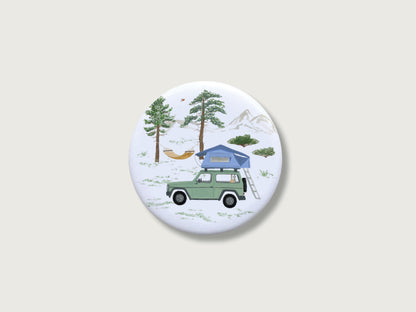 Runder Magnet mit handillustriertem Camping-Motiv. Ein Jeep mit Dachzelt steht in der Natur. Im Hintergrund sind Bäume, Berge und eine gespannte Hängematte zu sehen. Camping-Idylle.