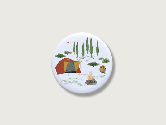 Runder Magnetbutton mit handillustriertem Camping-Motiv. Inmitten der Natur ist ein Zelt (Iglu) vor einem Lagerfeuer zu sehen.