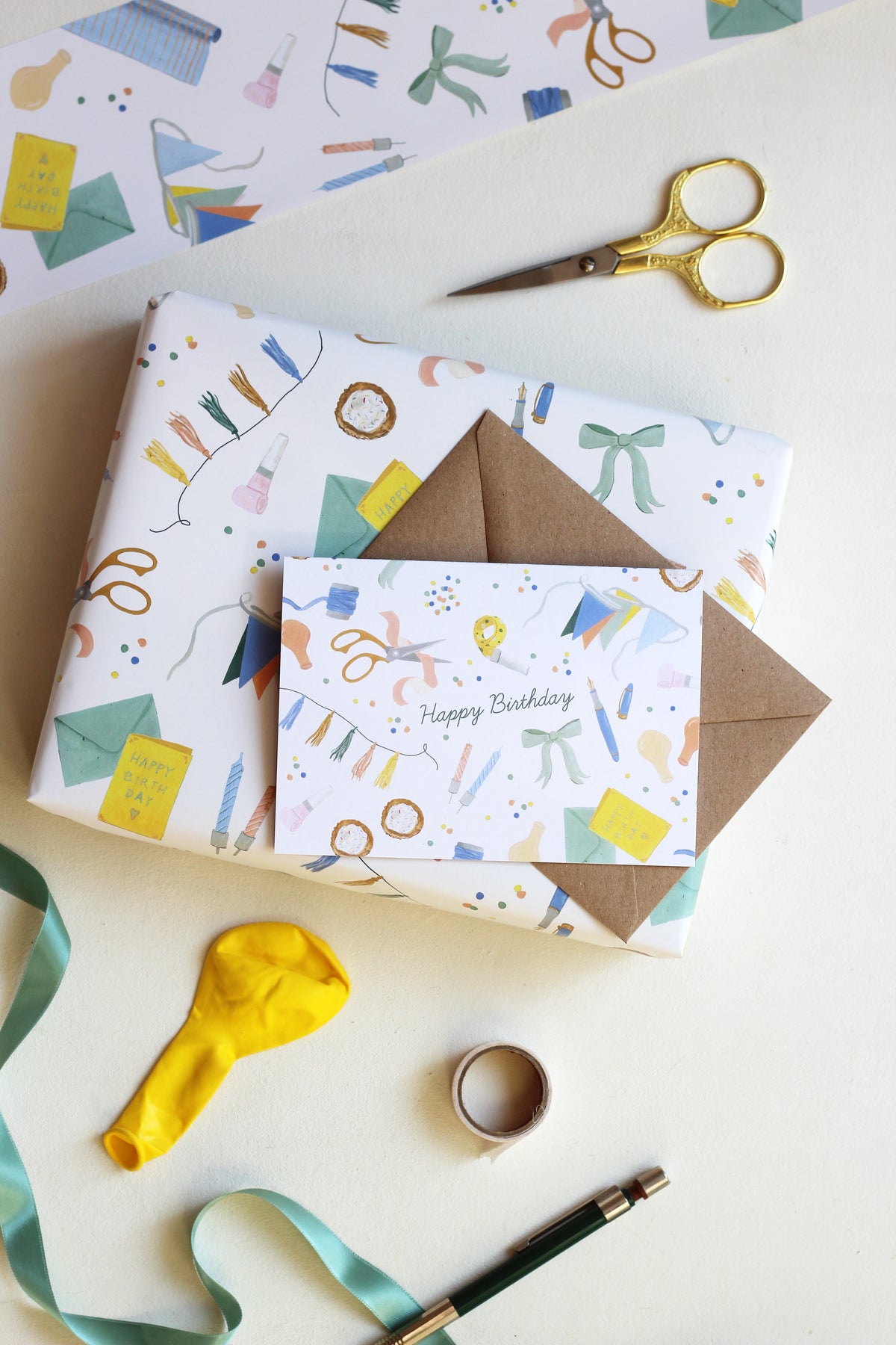 Das Bild zeigt einen hellen Hintergrund, auf dem ein eingepacktes Geschenk mit dazu passender Geburtstagskarte liegt. Grußkarteund Geschenkpapier zeigen eine handillustriertes Muster mit verschiedenen Partygegenständen, wie beispielsweise Girlande, Schleife, Luftballon und Kröte. Neben der Papeterie liegen noch aufgerollte Geschenkverpackung, Luftballon, Schere und Stift. 