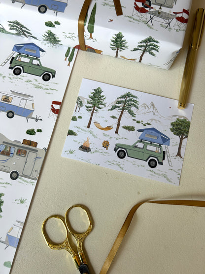 Kunstvolle Camping-Postkarte mit handgemalter Illustration eines Geländewagens mit Dachzelt, Lagerfeuer und Hängematte, arrangiert mit passendem Geschenkpapier.