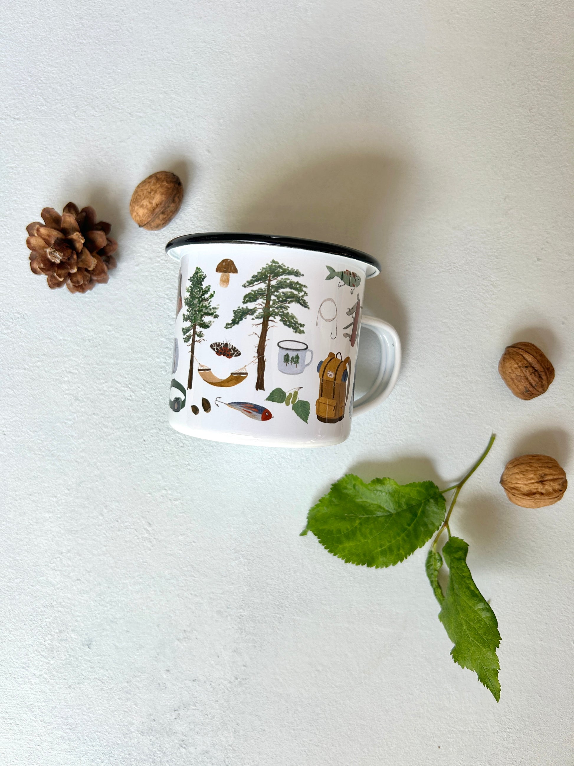 Eine weiße Emaille-Tasse mit schwarzem Rand steht im Zentrum einer Flatlay-Dekoration auf hellem Untergrund. Die Tasse ist mit feinen, handgemalten Outdoor- und Waldmotiven bedruckt, darunter Tannen, eine Hängematte, Bushcraft- und Survival-Werkzeuge und ein Pilz. Drumherum sind ein Tannenzapfen, Walnüsse und ein grüner Blätterzweig arrangiert.