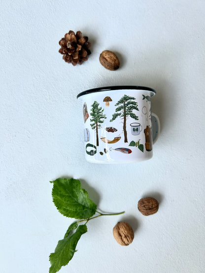 Eine weiße Emaille-Tasse mit schwarzem Rand steht im Zentrum einer Flatlay-Dekoration auf hellem Untergrund. Die Tasse ist mit feinen, handgemalten Outdoor- und Waldmotiven bedruckt, darunter Tannen, eine Hängematte und ein Pilz. Drumherum sind ein Tannenzapfen, Walnüsse und ein grüner Blätterzweig arrangiert.