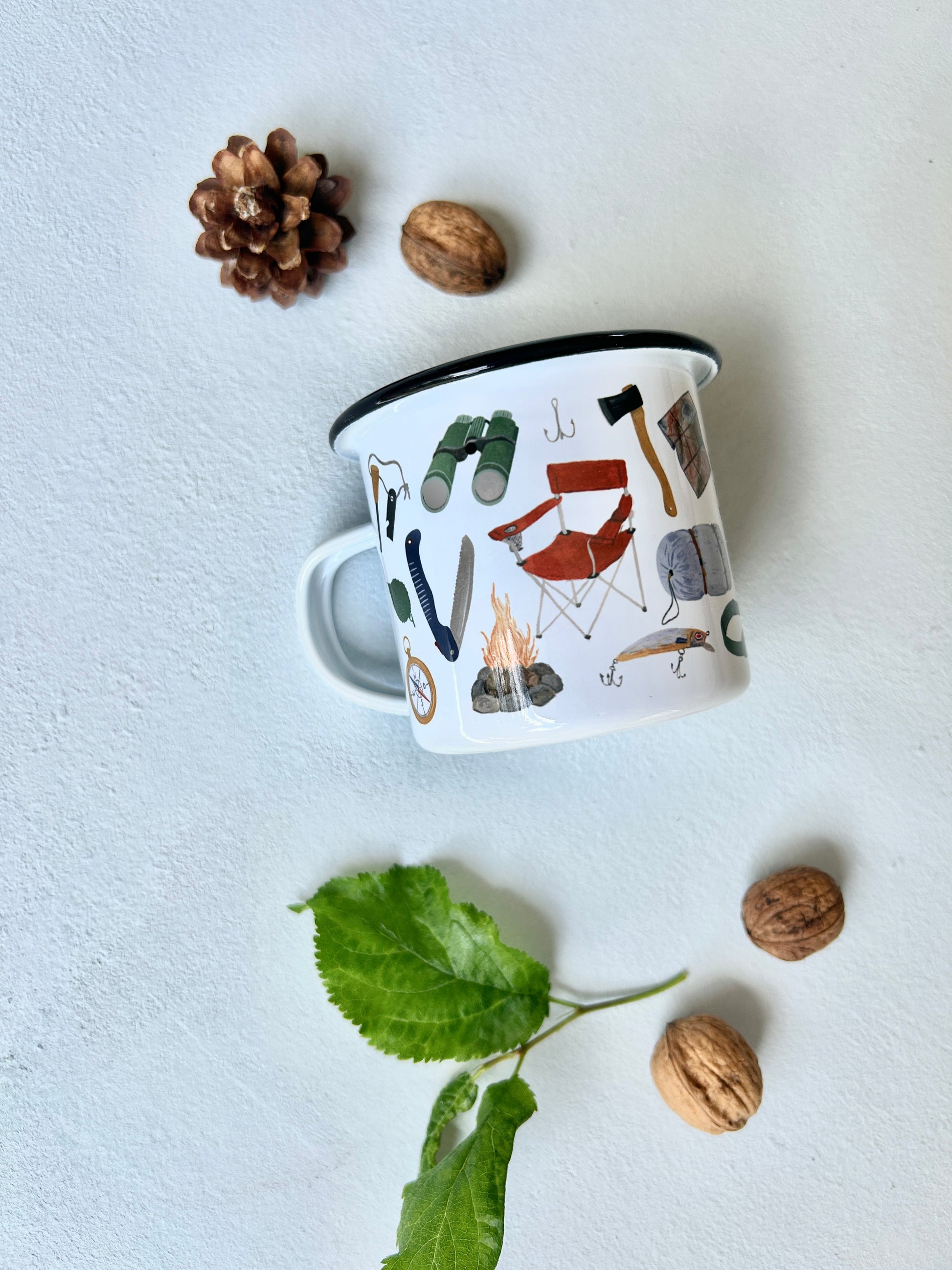Eine weiße Emaille-Tasse mit schwarzem Rand steht im Zentrum einer Flatlay-Dekoration auf hellem Untergrund. Die Tasse ist mit feinen, handgemalten Camping- und Survival-motiven bedruckt, darunter Schlafsack, Fernglas, Säge, Kompass, Axt, Feuerstahl. Eine Tasse für Bushcraft, Survival oder zum Wandern oder Wald-Abenteuer. 