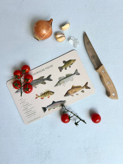 Berttchen mit handgemalten Illustrationen von heimischen Fische, das auf hellblauem Untergrund liegt. Daneben liegt ein Messer, sowie Zwiebeln und Knoblauch. Auf dem Brettchen liegen ein paar Cocktail-Tomaten. 