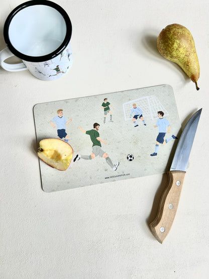 Ein graugrünes Frühstücksbrettchen mit handgemalten Illustrationen spielender Fußballer liegt auf einer hellen Tischplatte. Im Vordergrund schneidet ein Holzmesser ein Stück Apfel direkt auf dem Brettchen an. Links oben steht eine weiße Emaille-Tasse mit schwarzem Rand und passendem Fußball-Motiv, rechts daneben liegt eine frische Birne. Am unteren Rand des Brettchens ist die dezente Webadresse „www.HERZundPAPIER.com“ zu sehen.