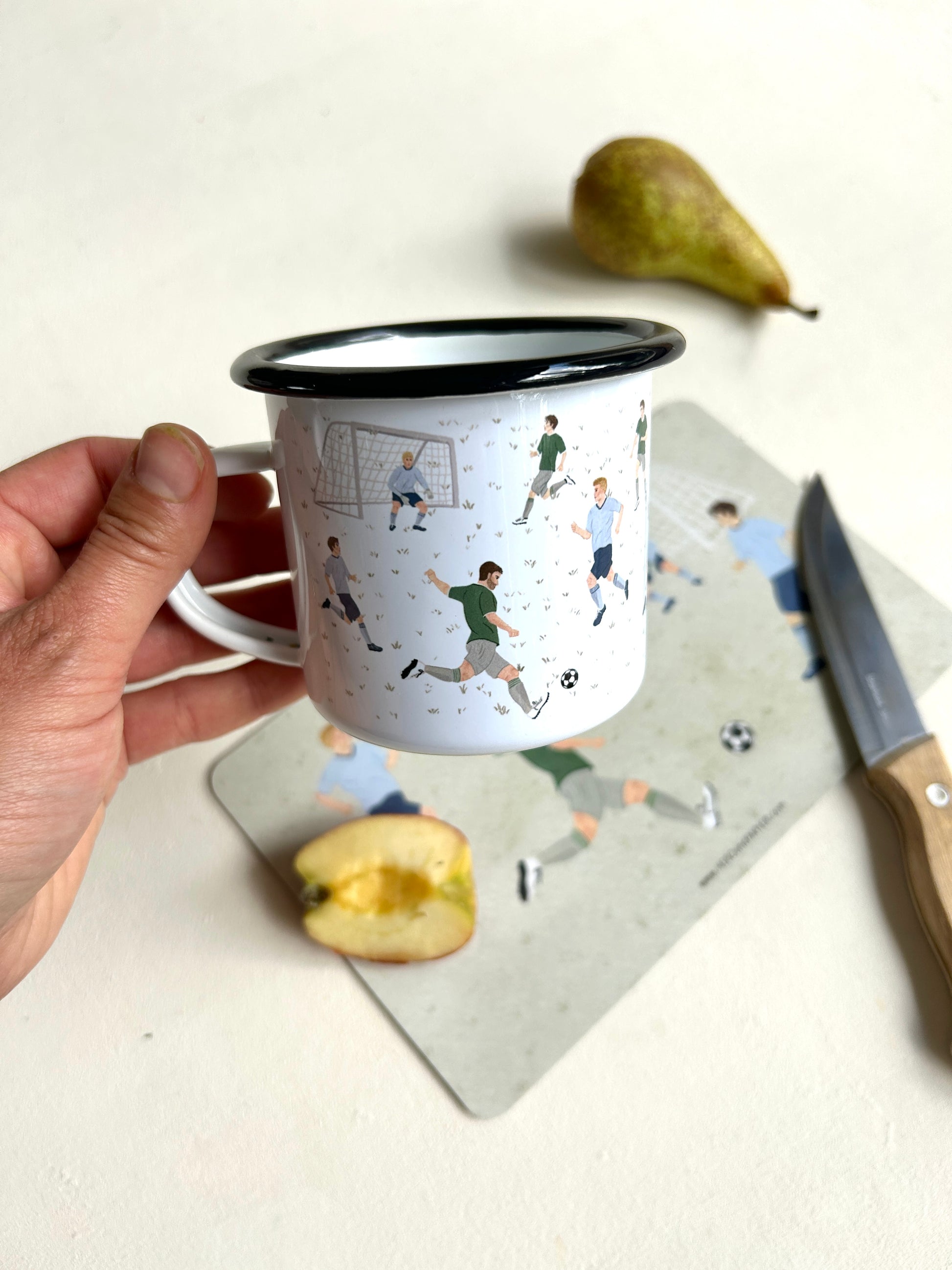 Eine Hand hält eine weiße Emaille-Tasse mit schwarzem Rand in die Kamera. Auf der Tasse sind feine Illustrationen von Fußballspielern in hellblauen und grünen Trikots sowie ein Torwart im Tor zu sehen. Im unscharfen Hintergrund liegen das passende graugrüne Fußball-Frühstücksbrettchen, eine halbe Apfelspalte, ein Küchenmesser mit Holzgriff und eine reife Birne auf hellem Untergrund.