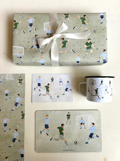 Flachgelegte Produktübersicht einer Fußball-Kollektion aus der Vogelperspektive auf weißem Grund. Oben liegt ein großes, mit einer weißen Seifenschleife verpacktes Geschenk in graugrünem Fußball-Geschenkpapier. Darunter sind eine Postkarte mit spielenden Fußballern, eine weiße Emaille-Tasse, ein passendes Frühstücksbrettchen sowie ein Ausschnitt des Geschenkpapier-Bogens harmonisch nebeneinander arrangiert. Die handgemalten Motive zeigen dynamische Fußball-Szenen.