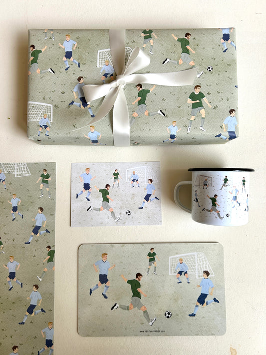 Flachgelegte Produktübersicht einer Fußball-Kollektion aus der Vogelperspektive auf weißem Grund. Oben liegt ein großes, mit einer weißen Seifenschleife verpacktes Geschenk in graugrünem Fußball-Geschenkpapier. Darunter sind eine Postkarte mit spielenden Fußballern, eine weiße Emaille-Tasse, ein passendes Frühstücksbrettchen sowie ein Ausschnitt des Geschenkpapier-Bogens harmonisch nebeneinander arrangiert. Die handgemalten Motive zeigen dynamische Fußball-Szenen.