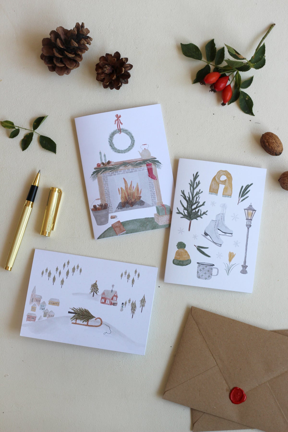 Drei handillustrierte Weihnachtskartenauf einem Tisch mit passenden Umschlägen neben weihnachtlicher Natur-Deko. Die Klappkarten zu Weihnachten zeigen stimmungsvolle Illustrationen von einer gemütlichen Szene zu Hause, einer winterlichen Collage und einer Weihnachtlichen Schneelandschaft. 