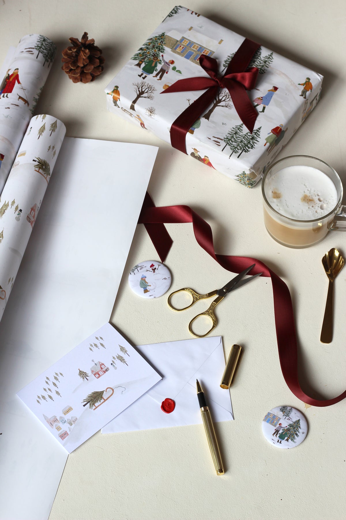 Auf einem hellen Hintergrund liegen zwei Bögen Weihnachtsgeschenkpapier. Schräg darüber ist ein Geschenk mit illustrierter Weihnachtslandschaft zu sehen. Darunter liegen Magnet-Buttons mit winterlichen und weihnachtlichen Motiven sowie eine Weihnachtskarte neben Füller, Schere, Geschenkband und Kaffee.