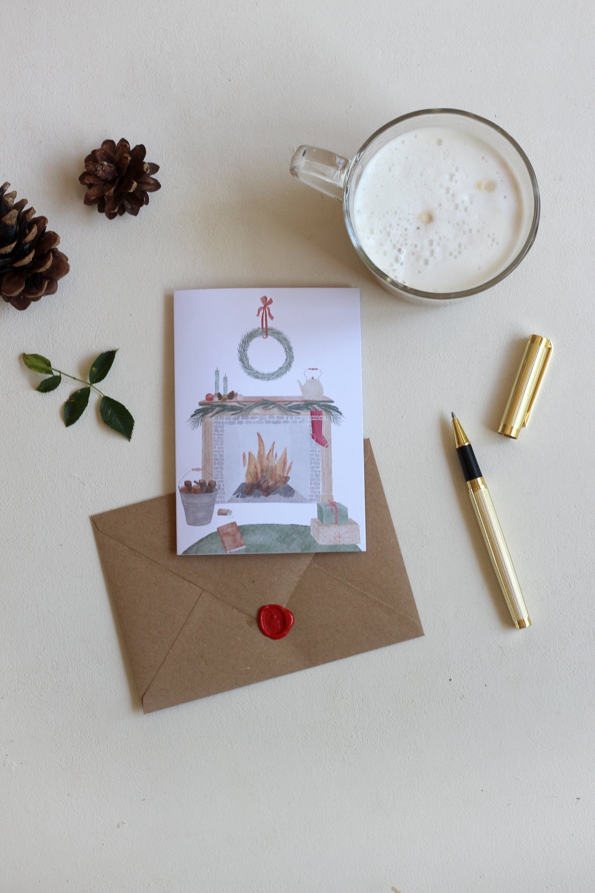 Weihnachtskarte mit handillustrierter , gemütlicher Szene von Zuhause. Das Motiv zeigt einen offenen Kamin, weihnachtlich dekoriert und davor ein Buch und Geschenke. Unter der Klappkarte liegt der passende Umschlag aus Kraftpapier. Daneben liegt ein Füller und steht eine Tasse Kaffee. 