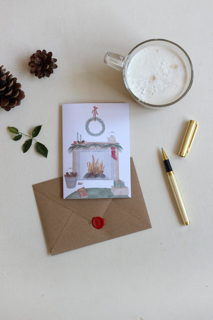 Weihnachtskarte mit handillustrierter , gemütlicher Szene von Zuhause. Das Motiv zeigt einen offenen Kamin, weihnachtlich dekoriert und davor ein Buch und Geschenke. Unter der Klappkarte liegt der passende Umschlag aus Kraftpapier. Daneben liegt ein Füller und steht eine Tasse Kaffee. 